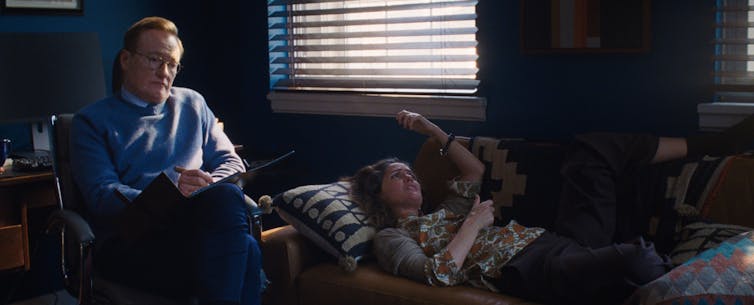 A man with red hair and glasses looks pained as a woman lies on a couch speaking.