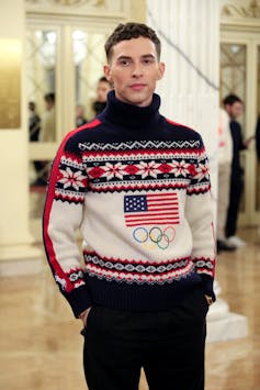 A young man, former figure skater Adam Rippon, stands with his hands in his pockets wearing an Olympics-themed sweater.