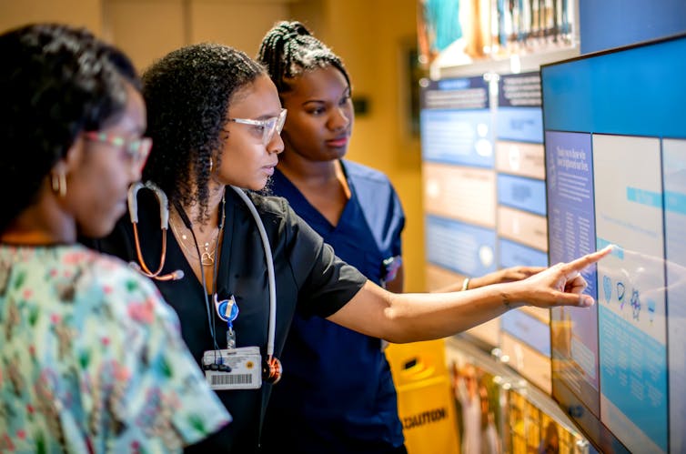 A group of Black nurses gather around a smartboard patient chart.