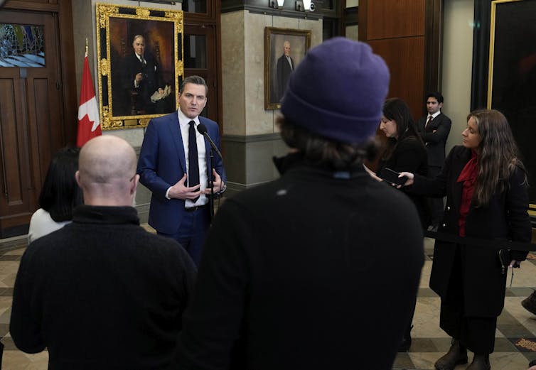 A man in a dark suit speaking in a large space, with other people seen from behind