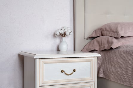 A set of white bedside drawers with a small base of flowers on top, and a bed laid with pink sheets and pillowcases.