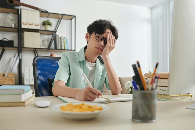 A teen boy wearing glasses sitting at a table with a notebook and pencil, resting his face on his hand