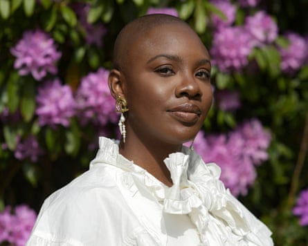 Yomi Adegoke wearing a white frilly blouse, standing in front of a green bush with big pink/purple flowers