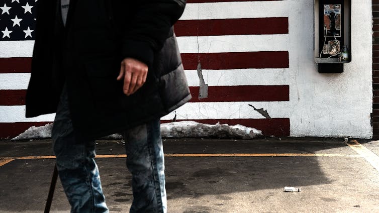 A person, visible torso down, walks through an economically stressed urban area, with a mural of an American flag behind them.