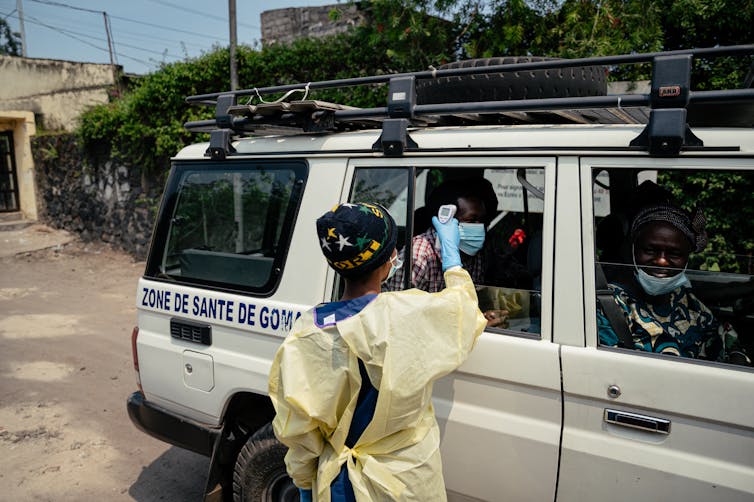 A person in protective gear checks the temperature of people in a white van.