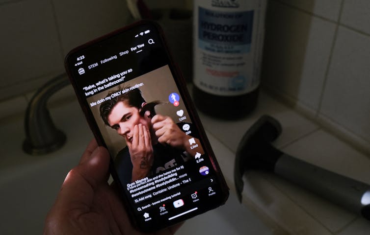 A hand holding a mobile phone on which a video has a young man smashing his face with a hammer.