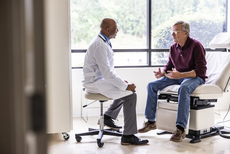 An older male doctor and older male patient talk in a medical exam room.