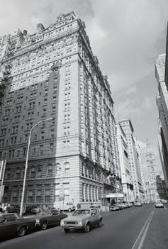 Black-and-white photo of large ornate building alongside two-way street with 1970s-era cars