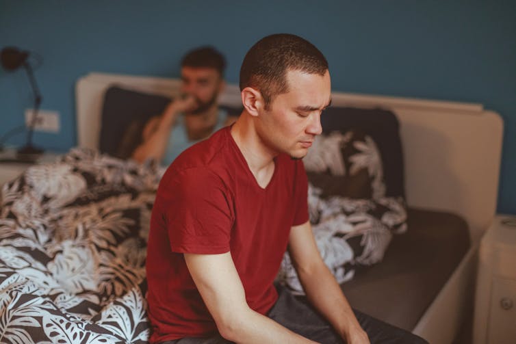 Person sitting on the edge of a bed, facing away from another person sitting against the headboard