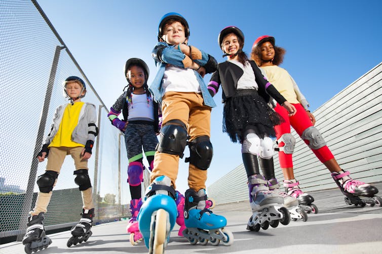 Five children on rollerblades wearing helmets and other safety gear pose in a skate park