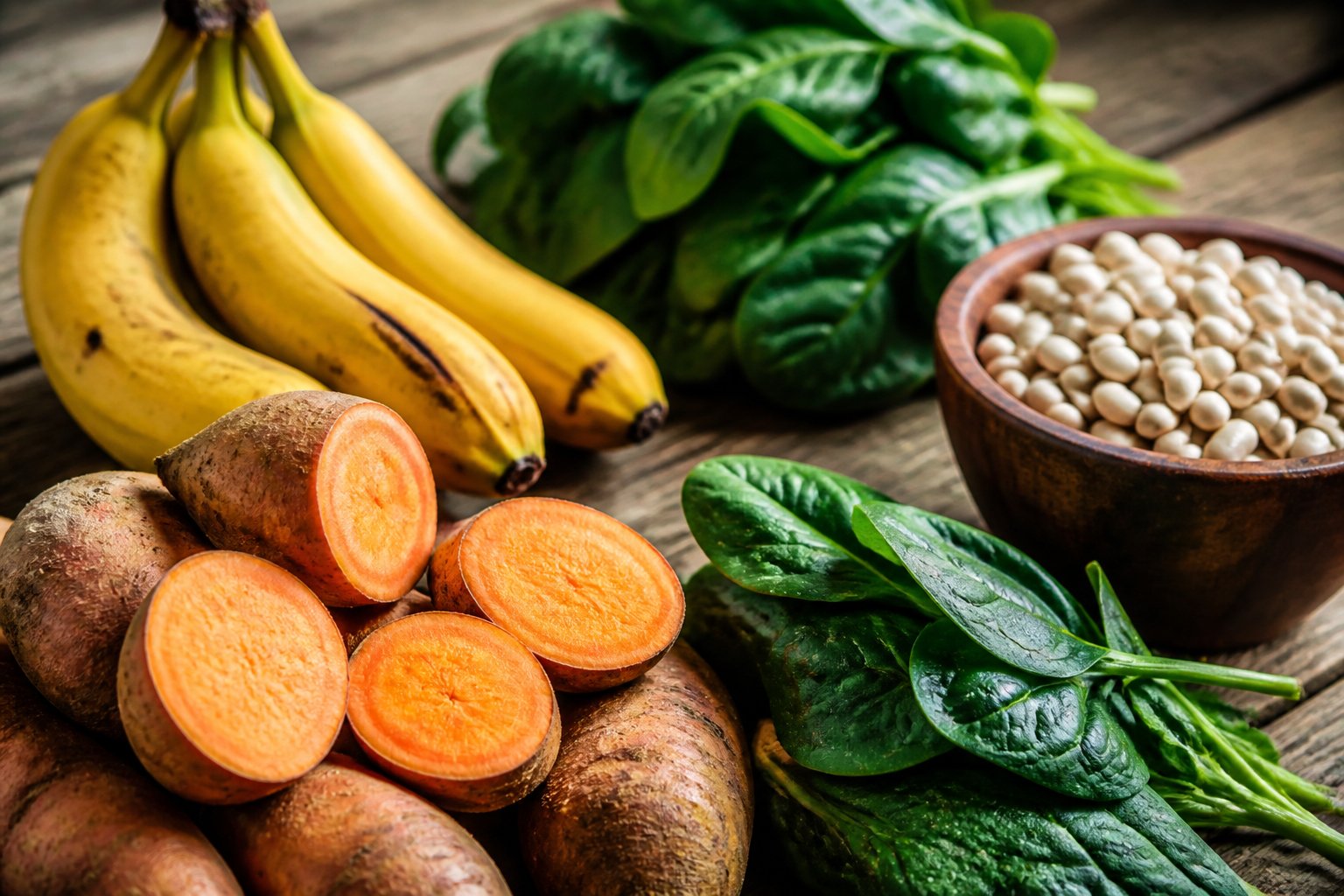 Close-up of potassium-rich foods including bananas, sweet potatoes, spinach, and white beans arranged on a wooden table.