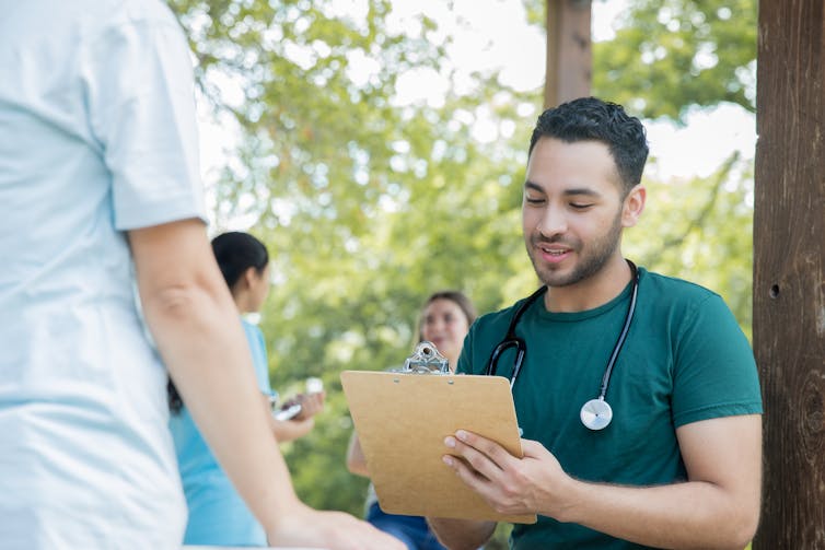 A male doctor or nurse with a stethoscope around his neck writes on a clipboard while talking to someone at a community health event