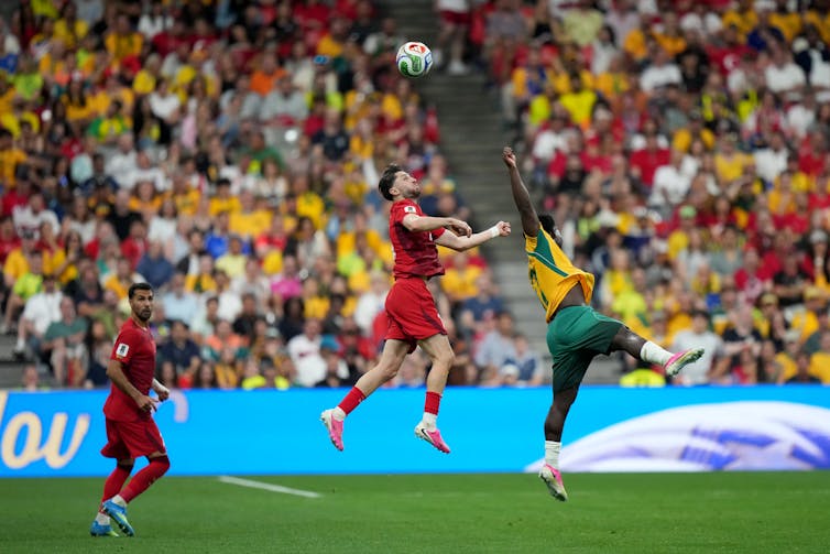 Two soccer players in mid-air with a soccer ball in flight, with crowded stands in the background
