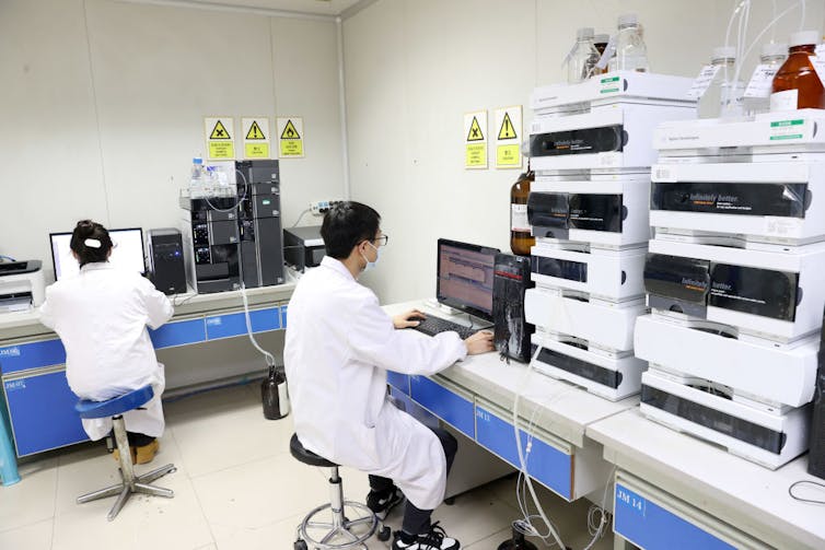 Two lab workers in white coats sitting at computers beside stacks of lab equipment and machines