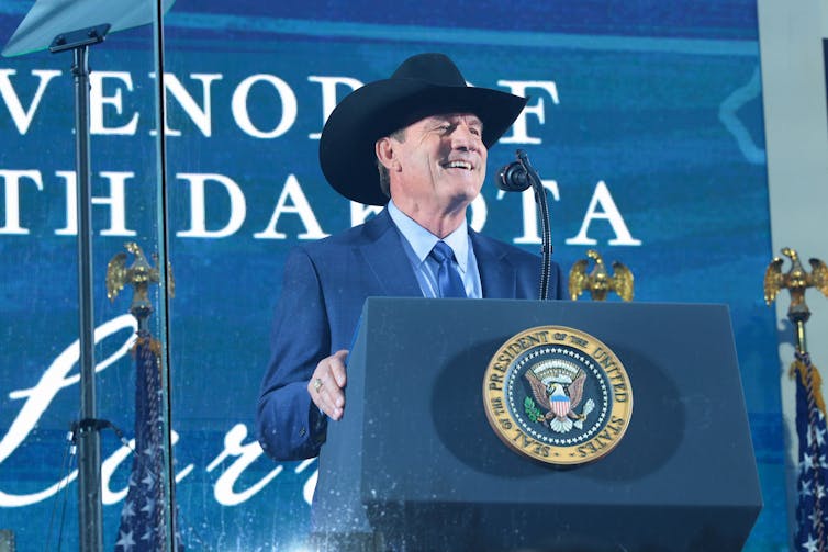 A man in a blue suit and wearing a cowboy hat gives a speech.