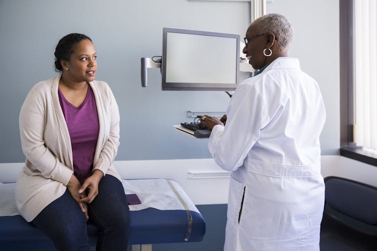 Patient sitting on exam room table talking to doctor, who is standing up at computer