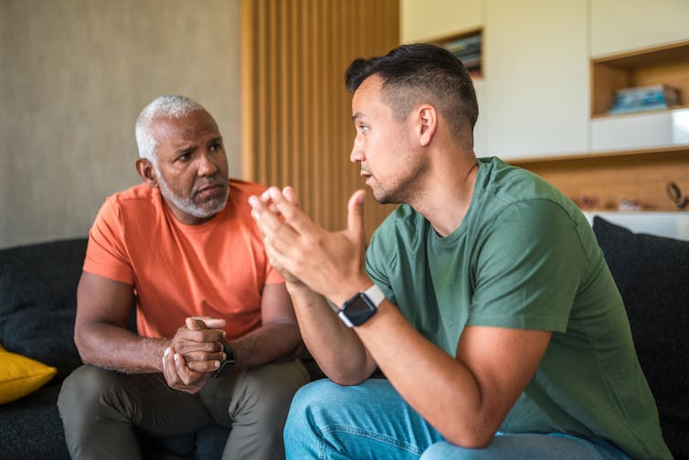 Two middle-aged men of diverse racial backgrounds engage in an intimate conversation while sitting in a living room.