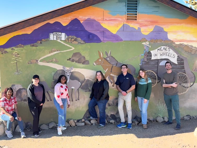 Seven people stand in front of a colorful mural.