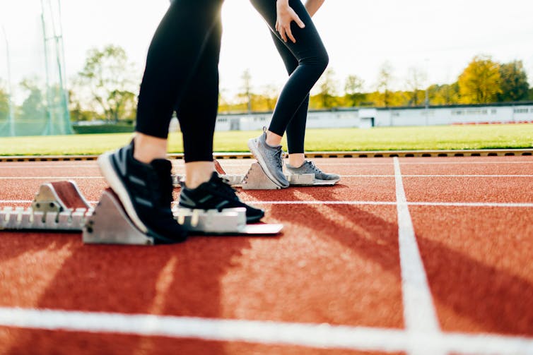 The calves of two runners wearing black leggings are visible on a red track, as the runners get into position with starting blocks.