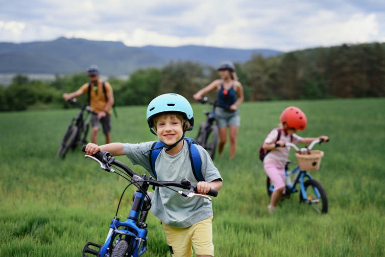 A child in a blue bike helmet with a blue bicycle in the foreground, with two adults on bikes and another child in a red helmet on a bike in the background in a grassy area
