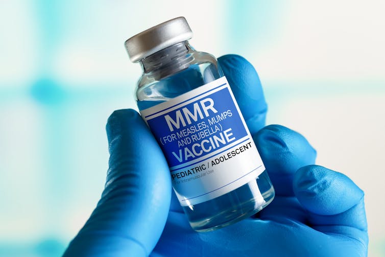 A gloved hand holding a bottle of MMR vaccine