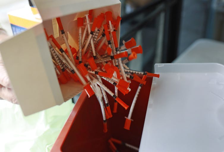 A white box releases many needles with an orange backing into a red square container.