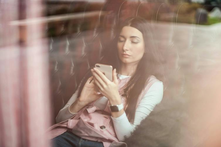 A woman seen through a window, seated and looking at a smartphone