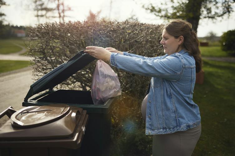 A pregnant woman throws a garbage bag into a trash can in a backyard.