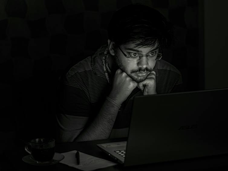 Black and white photo of a man in dark room, his face illuminated by the glow of a computer screen