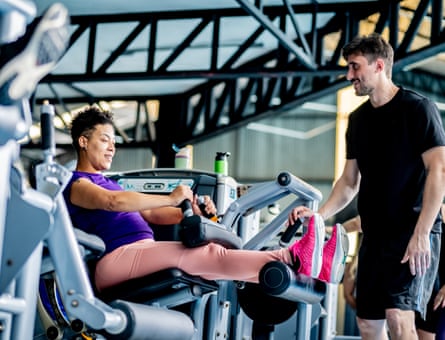 Mature woman does hamstring exercises on a machine with the help of a trainer at the gym.