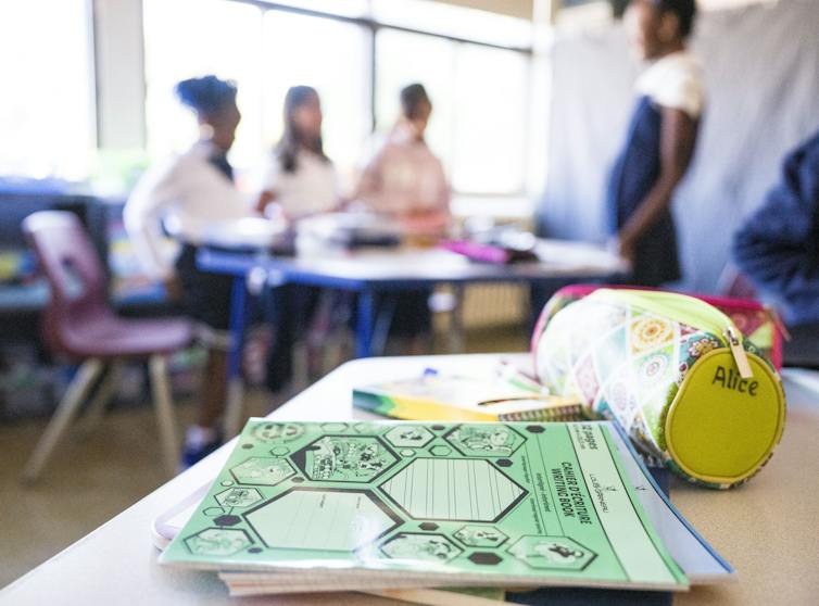 School supplies sitting on a desk while in the background the blurred images of students are seen congregating around a desk