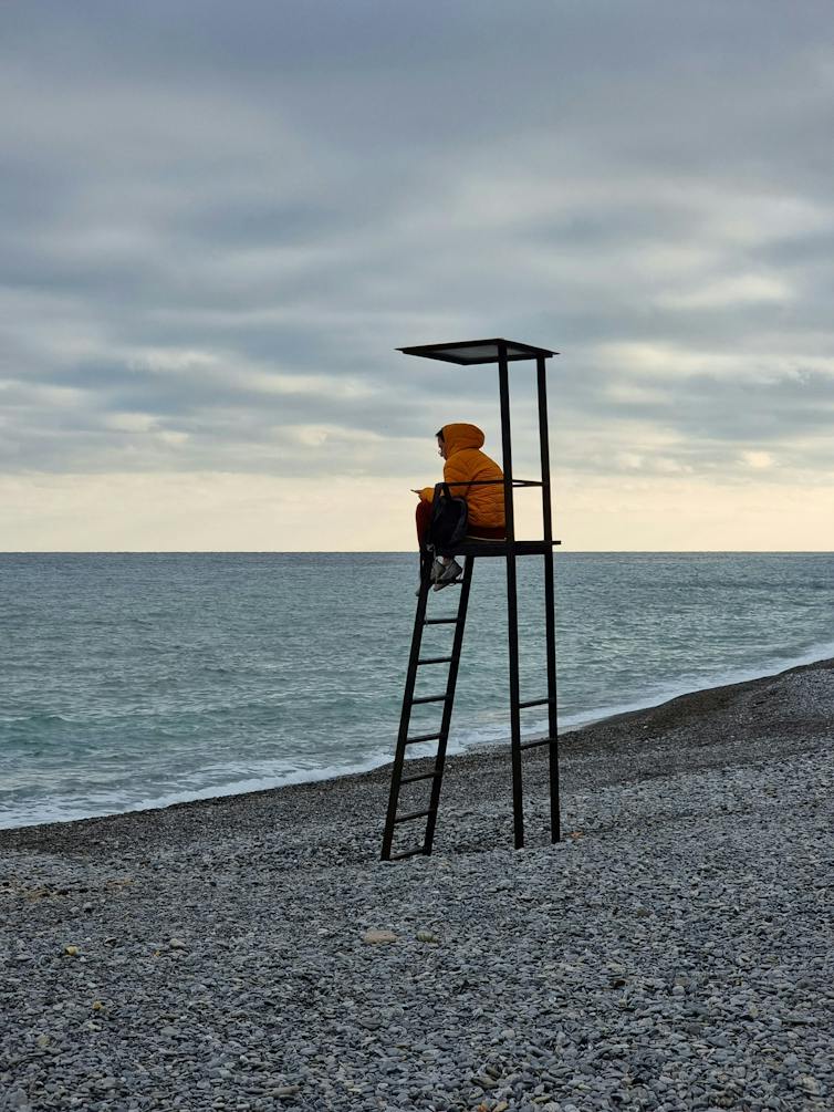 A lone lifeguard with an orange windbreaker sits atop his station, looking at the sea under an overcast sky.