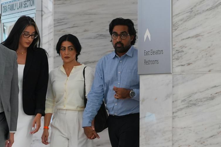 Two women and a man walk through a hallway, the man in a blue shirt holding hands with the woman in a white outfit.