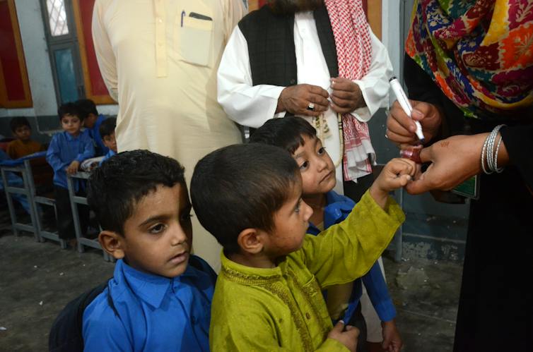 Three little boys standing together, one with his finger being marked by a woman